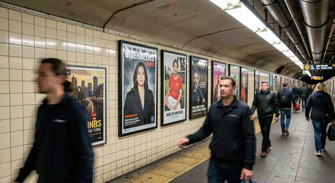 A New York subway platform with rows of framed advertisement posters along a tiled wall, commuters walking past in motion blur.
